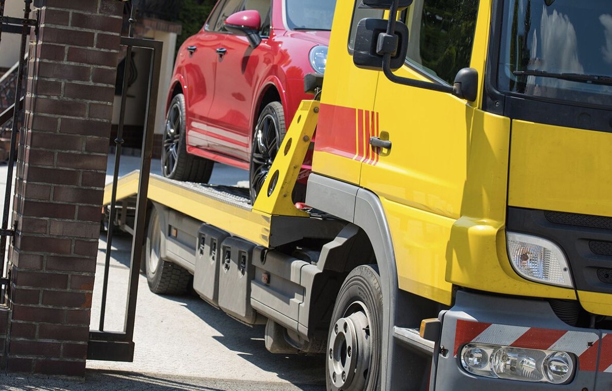 Yellow flatbed tow truck transporting a red compact car for a long distance tow in a residential area