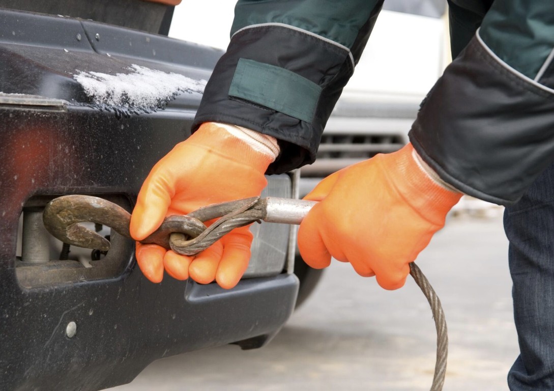 Tow truck operator in orange gloves attaching a heavy winch cable hook to a stuck vehicle bumper